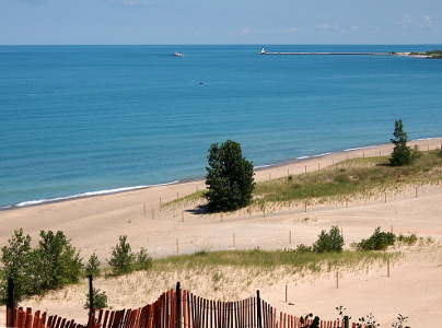 [The sand path to the shoreline is a switchback to make it less steep. Poles mark the path and there is vegetation growing outside of the path. In the far distance on the water is the lighthouse and a moving boat.]